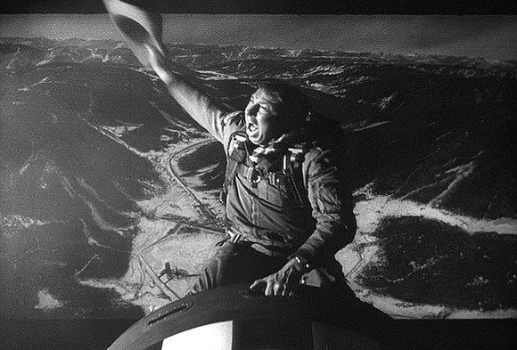 A man in a vintage astronaut suit joyfully waving a hat while sitting atop a bomb, with a mountainous landscape projected behind him.