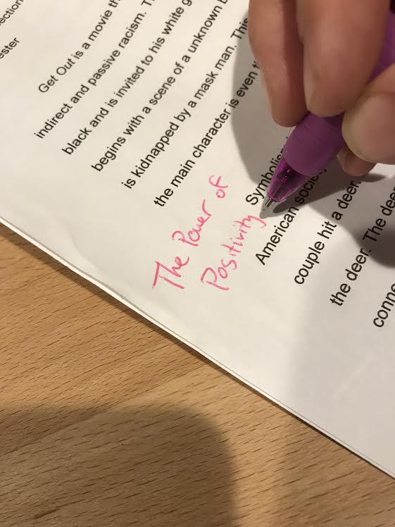 A hand holding a purple pen is marking a paper on a wooden surface. The words "The Power of Positivity" are highlighted in pink ink.