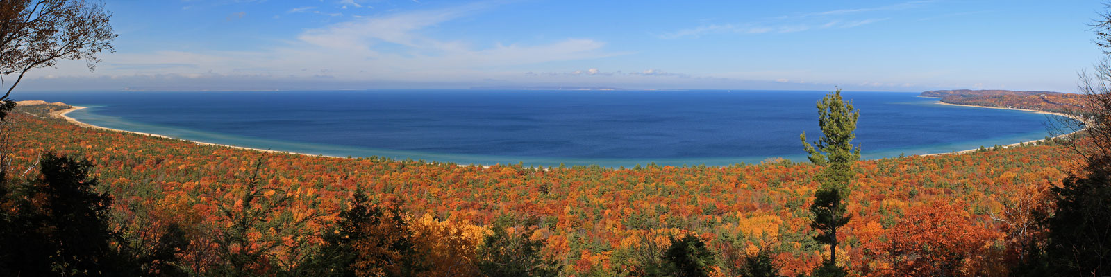 A panoramic view of Sleeping Bear Dunes National Park in Northern Michigan, showcasing vibrant autumn foliage along the shoreline and the blue waters of Lake Michigan.