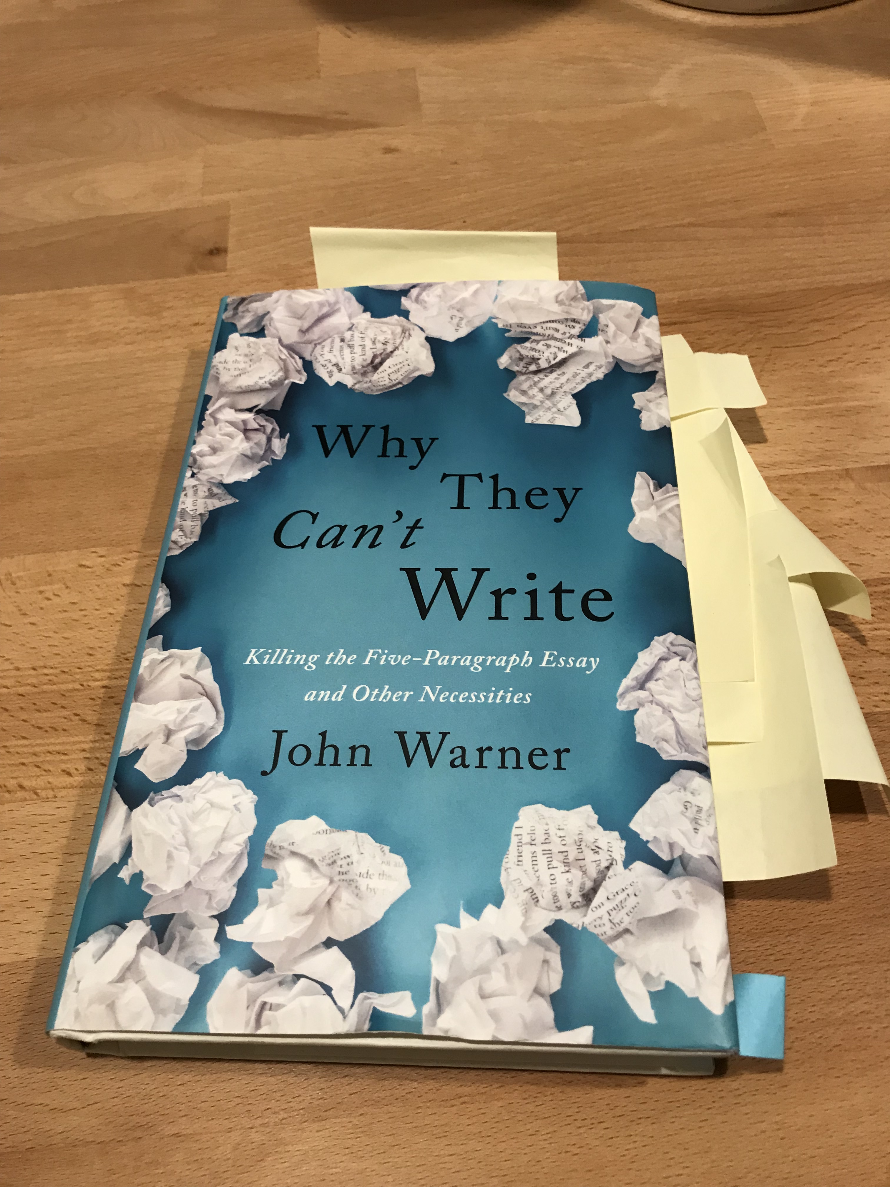 A book titled 'Why They Can't Write' by John Warner, featuring crumpled paper and tabs sticking out, lying on a wooden surface.
