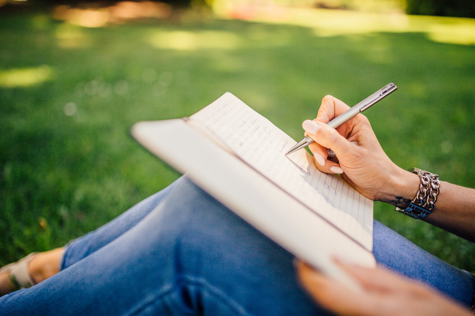 A close-up of a person's hand writing in a notebook while seated on grass, with a pen in hand and wearing a bracelet.