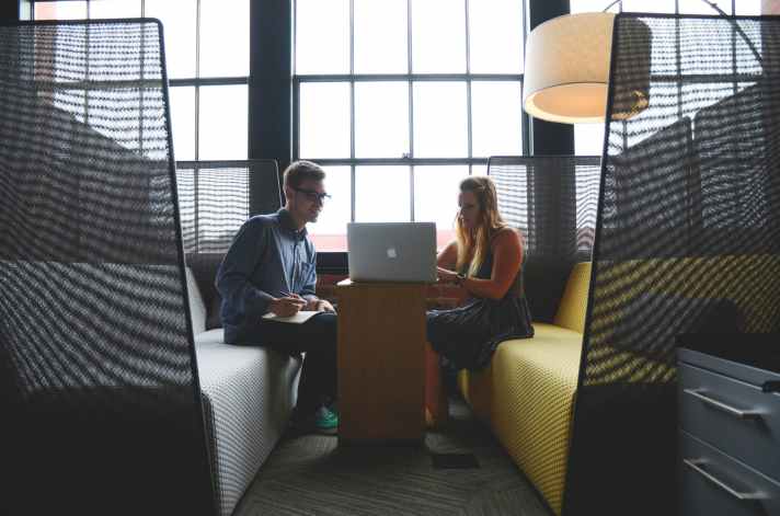 A man and a woman engage in a discussion while sitting at a table with a laptop in a modern workspace.