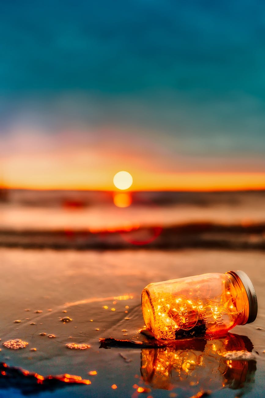 A glass jar with glowing lights inside, lying on a wet surface at the beach during sunset, with the sun setting on the horizon.