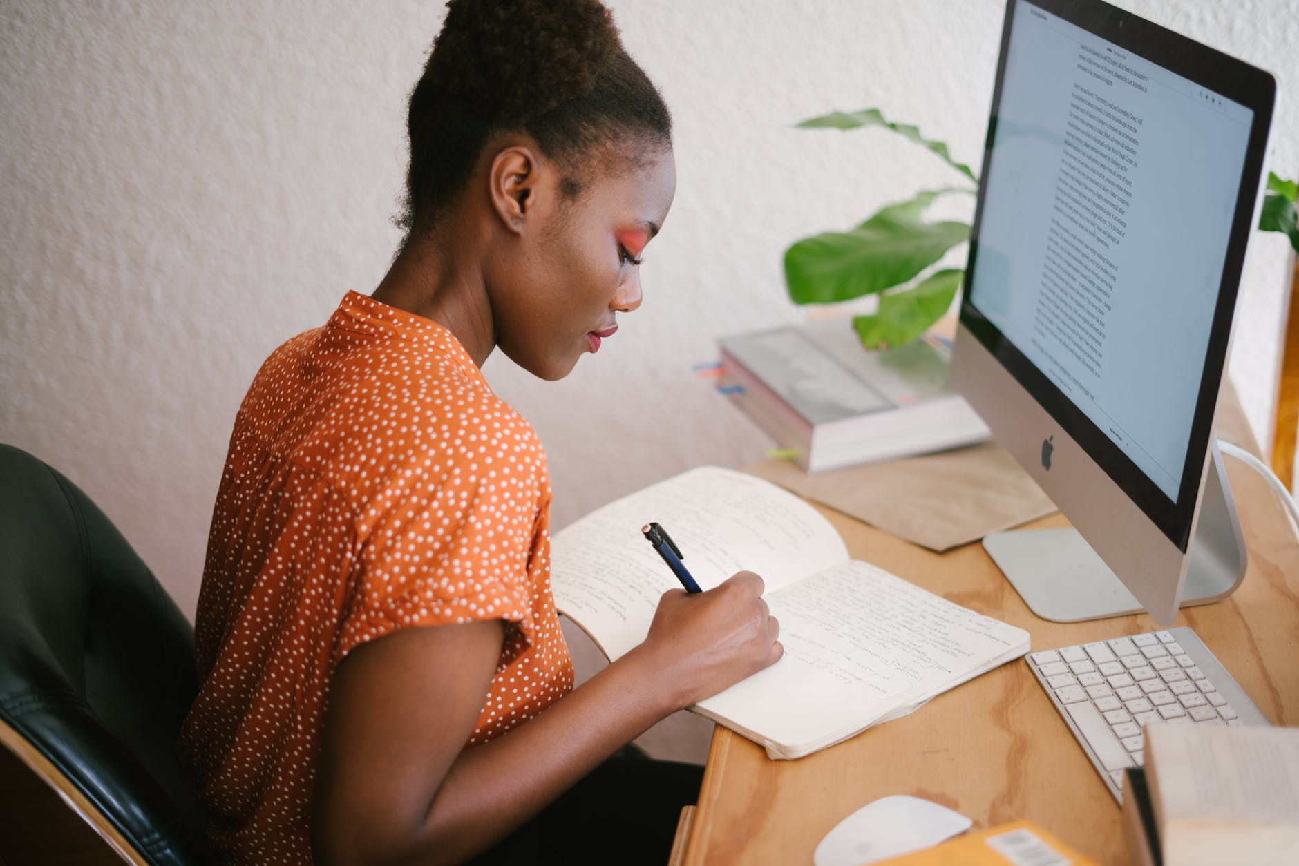 A person wearing an orange polka dot shirt is writing in a notebook at a wooden desk with a computer monitor and books in the background.