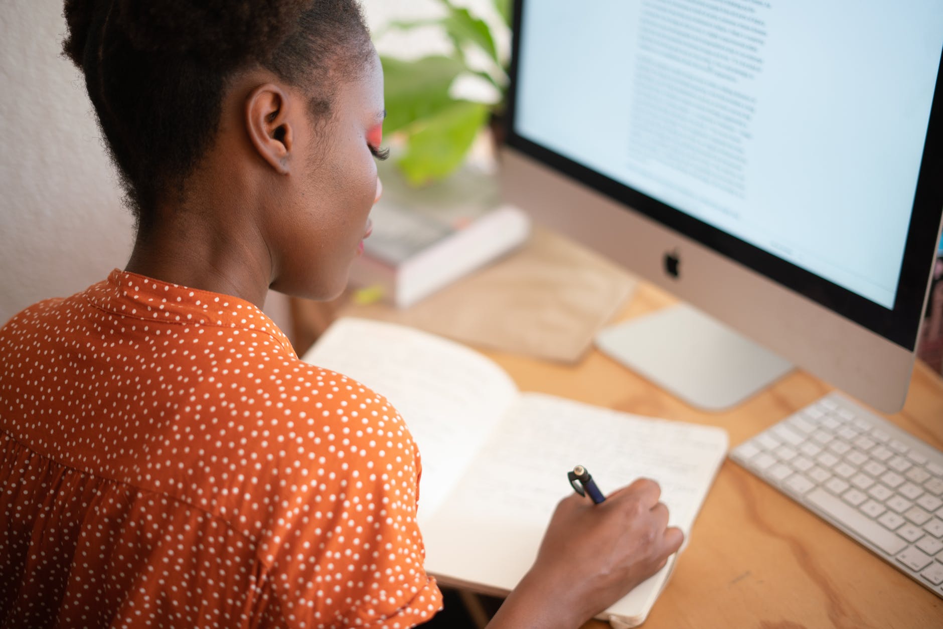 A person sitting at a desk, writing in a notebook while facing a computer screen.