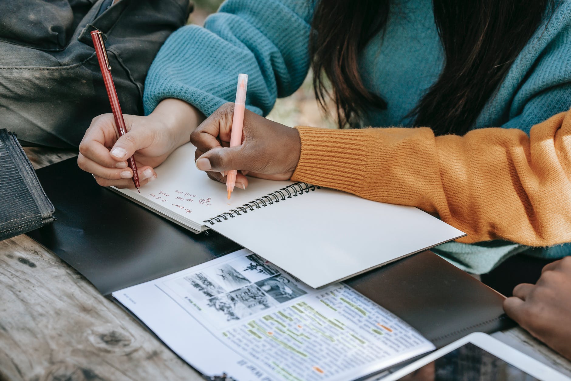Two people writing in a notebook, one using a red pen and the other a pink pen, while surrounded by papers and a black folder on a wooden table.