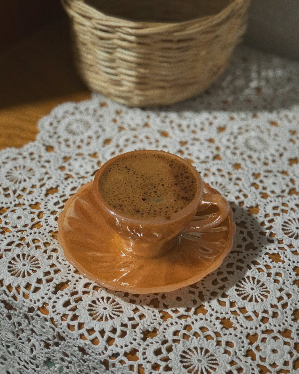 A small cup of coffee on a decorative lace tablecloth with a woven basket in the background.