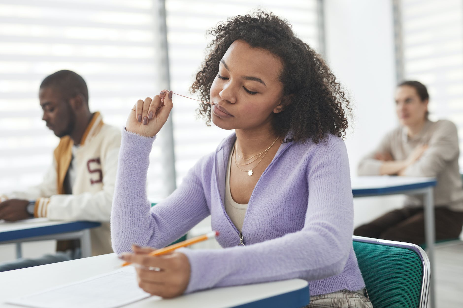 A student sitting at a desk in a classroom, thoughtfully holding a pencil while looking up, with two other students in the background focused on their work.