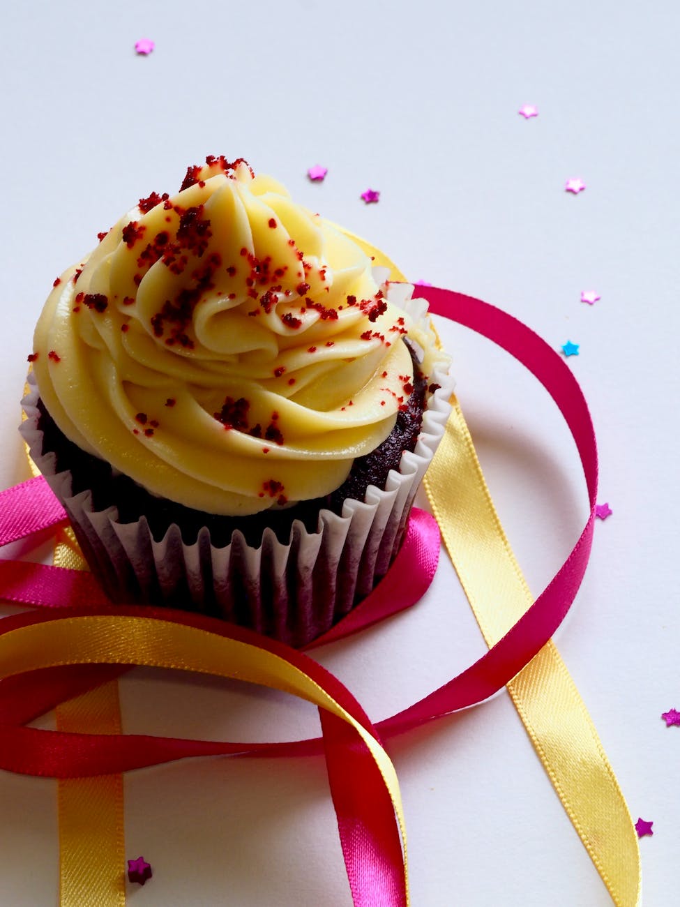 A close-up image of a chocolate cupcake topped with swirls of yellow frosting and red edible sprinkles, placed on a white surface with colorful ribbons and star-shaped confetti.