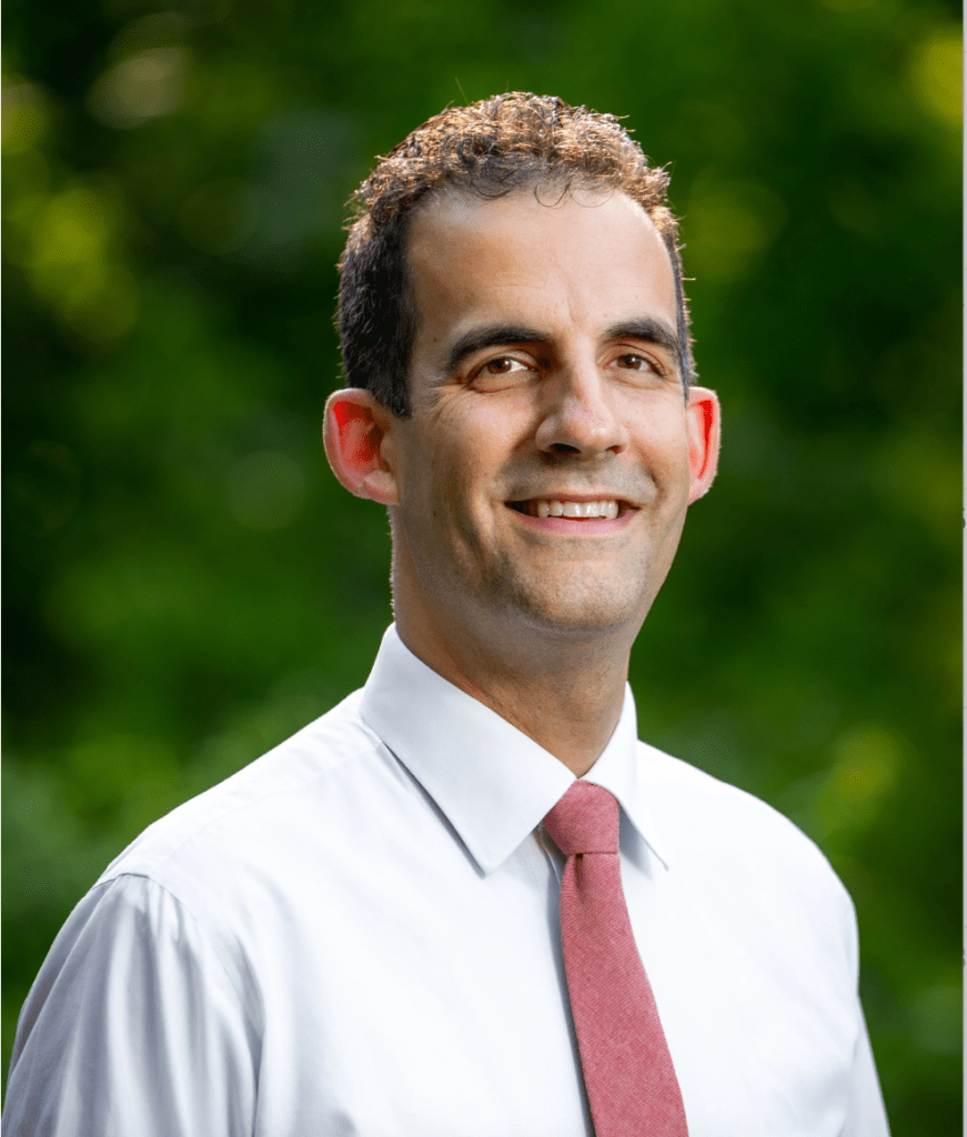 Brett Vogelsinger in a white shirt and pink tie smiles at the camera with a blurred green background.
