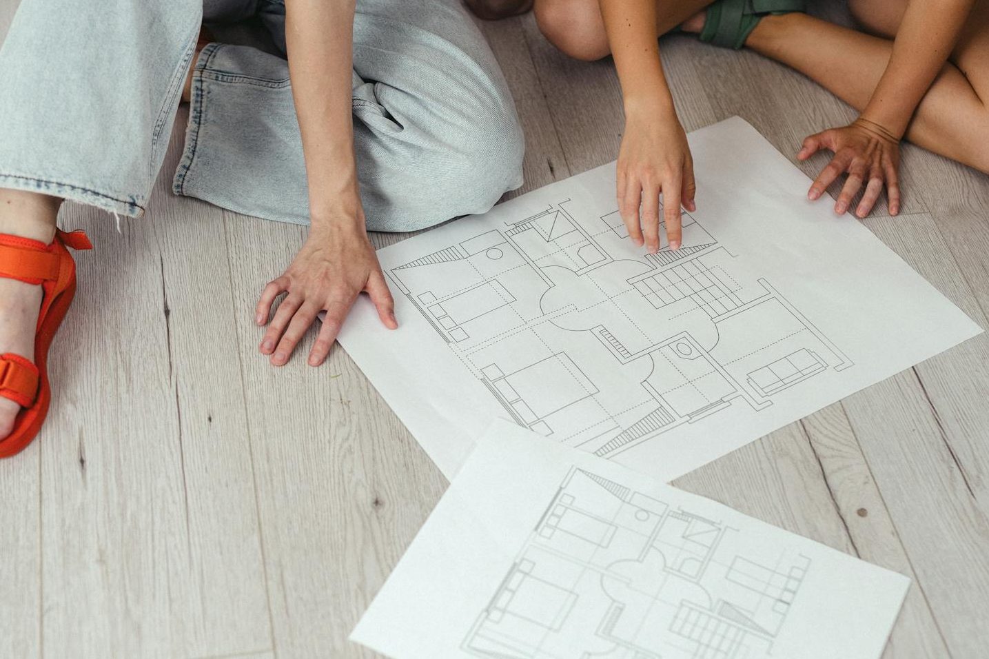 Two people examining house floor plans spread out on a light wooden surface.