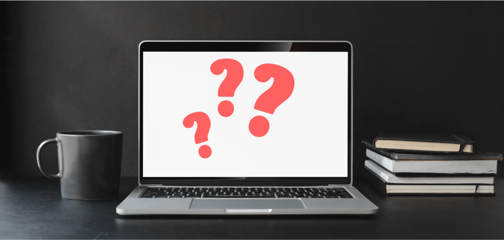 A laptop on a desk displaying three large red question marks on the screen, next to a coffee mug and a few stacked books.
